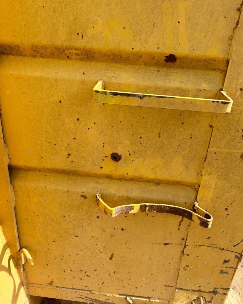 Close-up of a weathered yellow container wall with a small rusted hole between two metal rungs. The lower rung is crushed and bent.