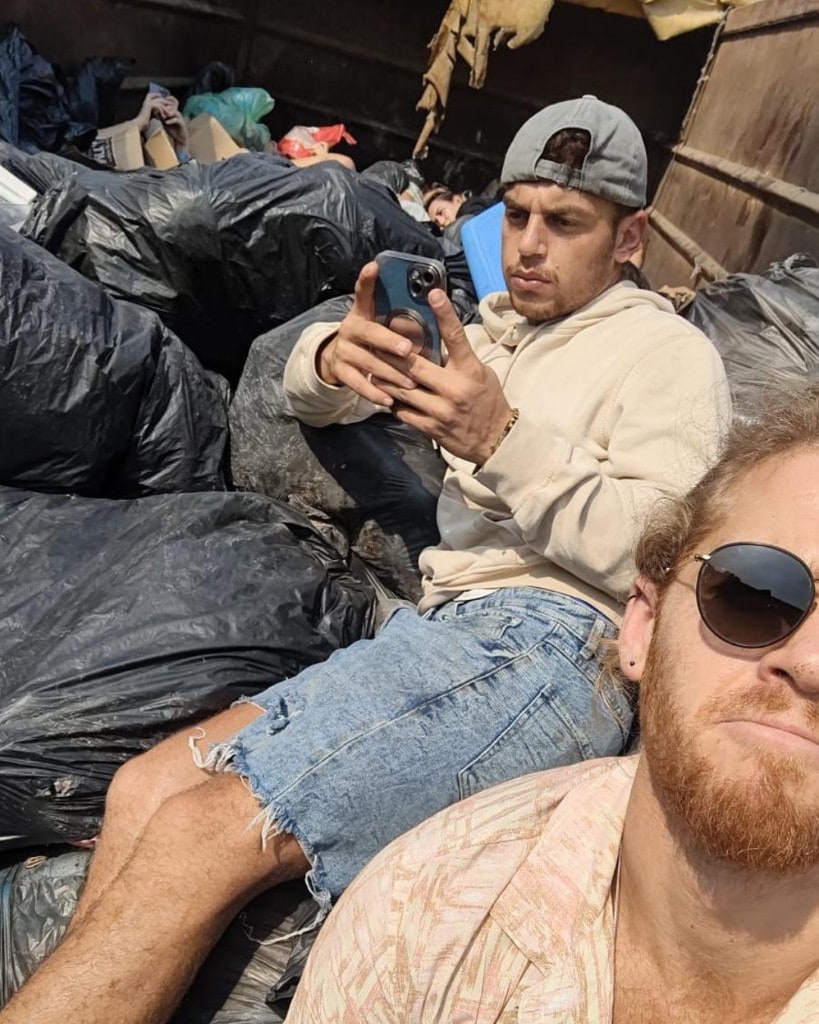 Young people sit among black refuse bags inside a dark metal container. One person in a cream hoodie looks at a phone; another person wearing sunglasses is in the foreground.