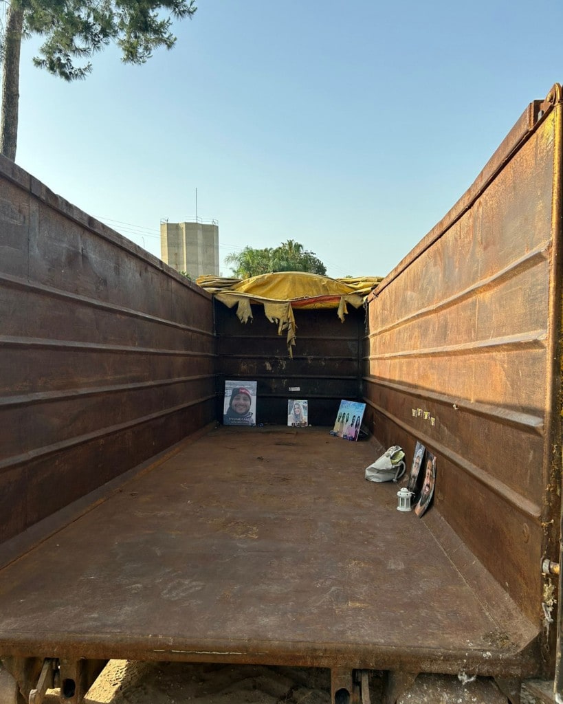 Looking into the rusted interior of a metal container. Framed portraits, a small lantern and a tattered yellow tarp stand inside, with sky and trees visible above.