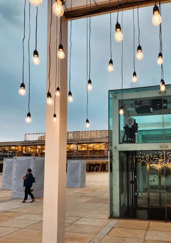 Twilight at Habima Square with hanging memorial bulbs in the foreground and a glowing Bring Them Home sign on the auditorium building