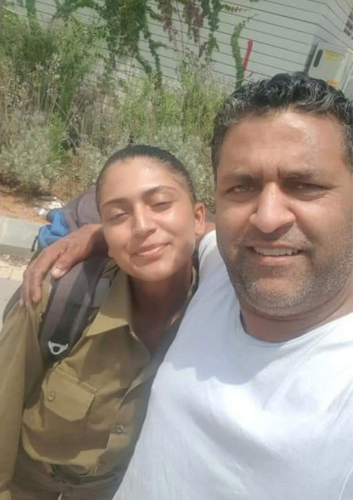 Kim in olive-green military uniform with a large backpack, smiling with her eyes closed as a man puts his arm around her outdoors